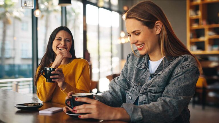Two,Women,Laughing,Inside,A,Coffee,Shop.,Friends,Sitting,With