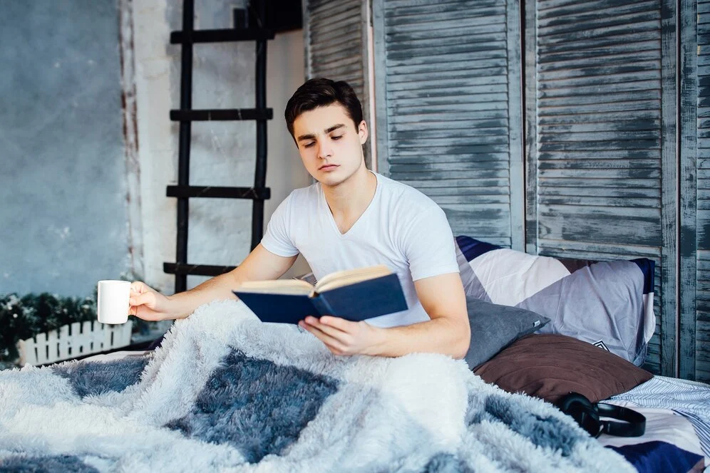 pretty-handsome-young-man-laying-shirtless-his-bed-holding-coffee-tea-cup-while-reading-book_496169-813