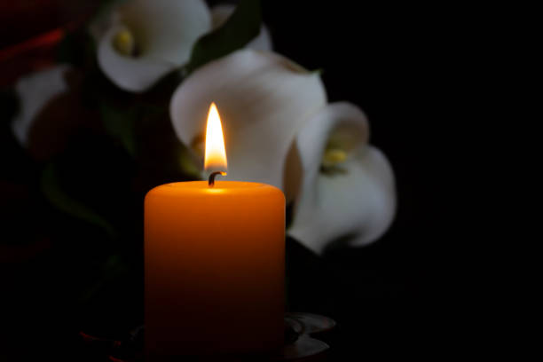 A close up of an orange candle and flame and lily flowers on a dark background.